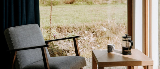 A french press on a table beside a chair and a window.