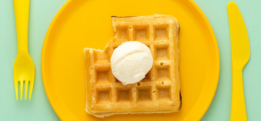 A waffle on a plate with a scoop of ice cream on top, and plastic cutlery.